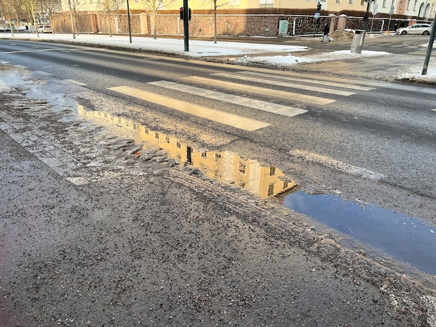 Kuva aurinkoisesta kadusta, jossa lumisulalätäkköön heijastuu pala keltaista kerrostaloa.