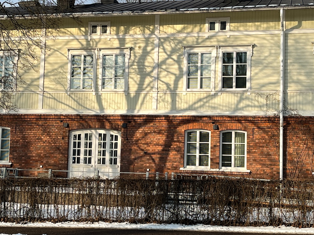 Puutalo, jonka seinälle heijastuu aurinkoa ja kahden lehdettömän puun varjot.