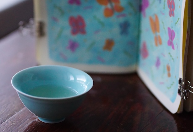 Pieni, turkoosi teekuppi, jossa on teetä, ja luonnoskirja, jossa kuva perhosista